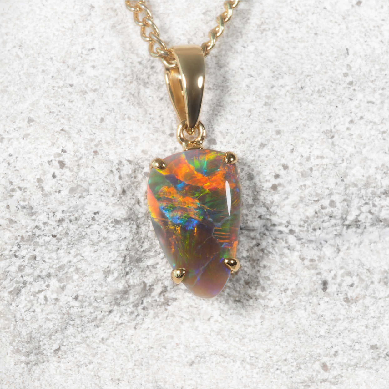 A gold opal necklace featuring a teardrop-shaped Lightning Ridge opal gemstone with vibrant colors including orange, green, blue, and yellow set on a textured light stone background.