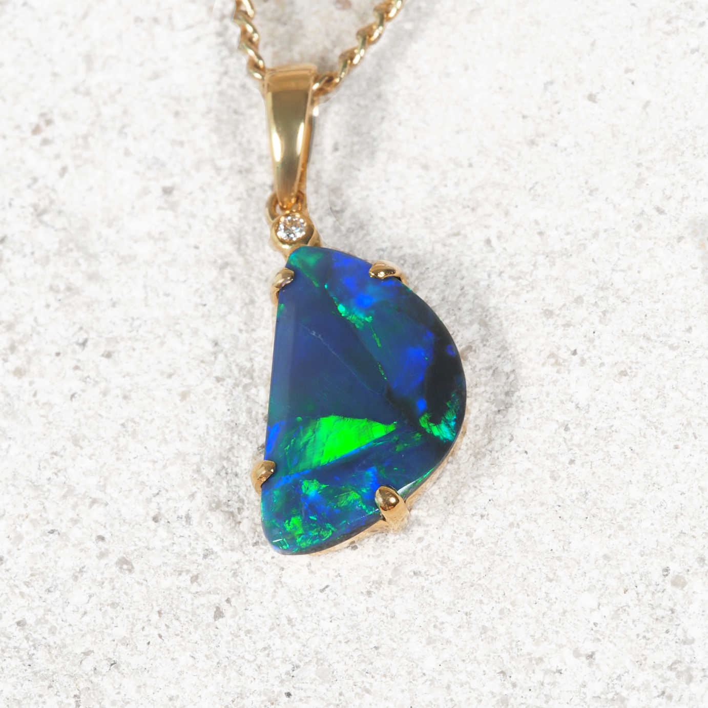 A gold black opal pendant necklace featuring a large, irregular-shaped opal gemstone with vibrant blue and green play-of-color, displayed on a textured light stone surface.