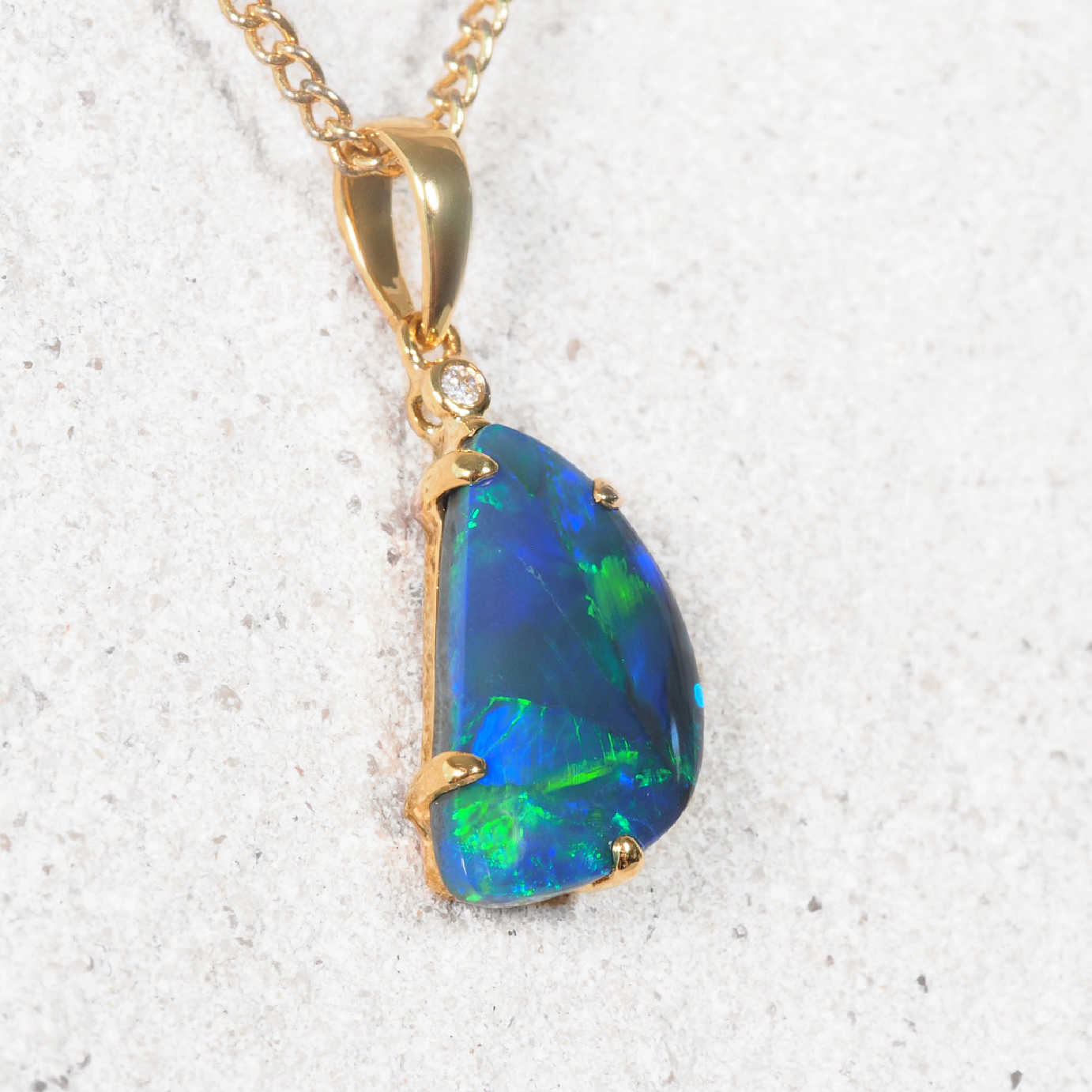 A authentic opal gold pendant necklace featuring a teardrop-shaped blue and green opal stone, displayed on a textured light gray surface.