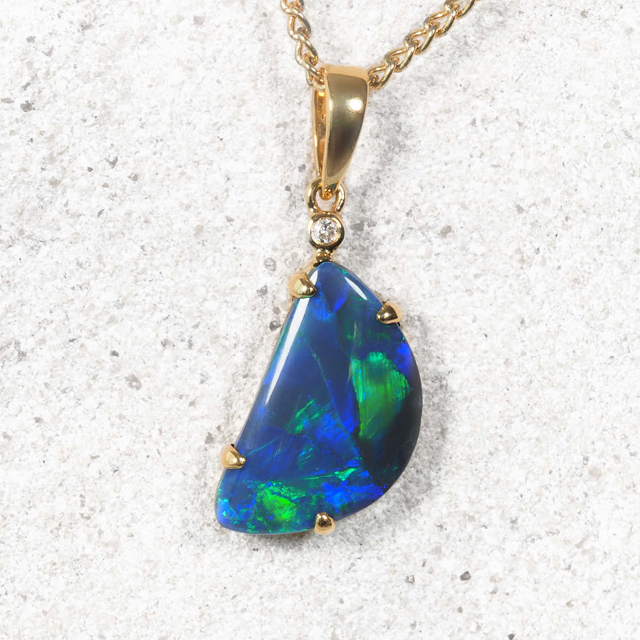 A gold Australian black opal pendant necklace featuring a large, irregular-shaped blue and green opal stone, accented with a small clear gemstone and set in gold, hanging from a gold chain against a light gray textured background.