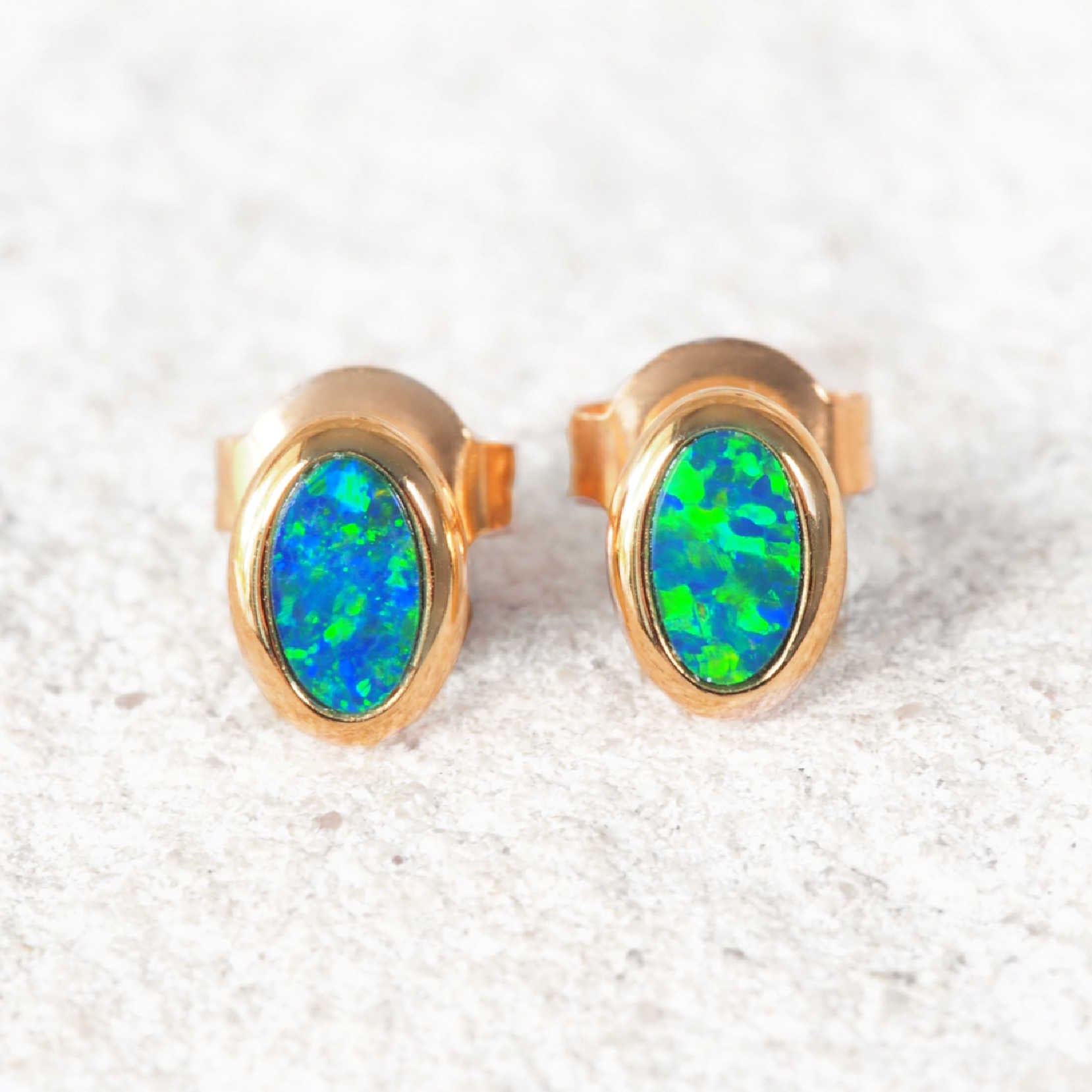 A pair of gold Australian opal stud earrings with oval green and blue opal gemstones, displayed on a textured light gray surface.