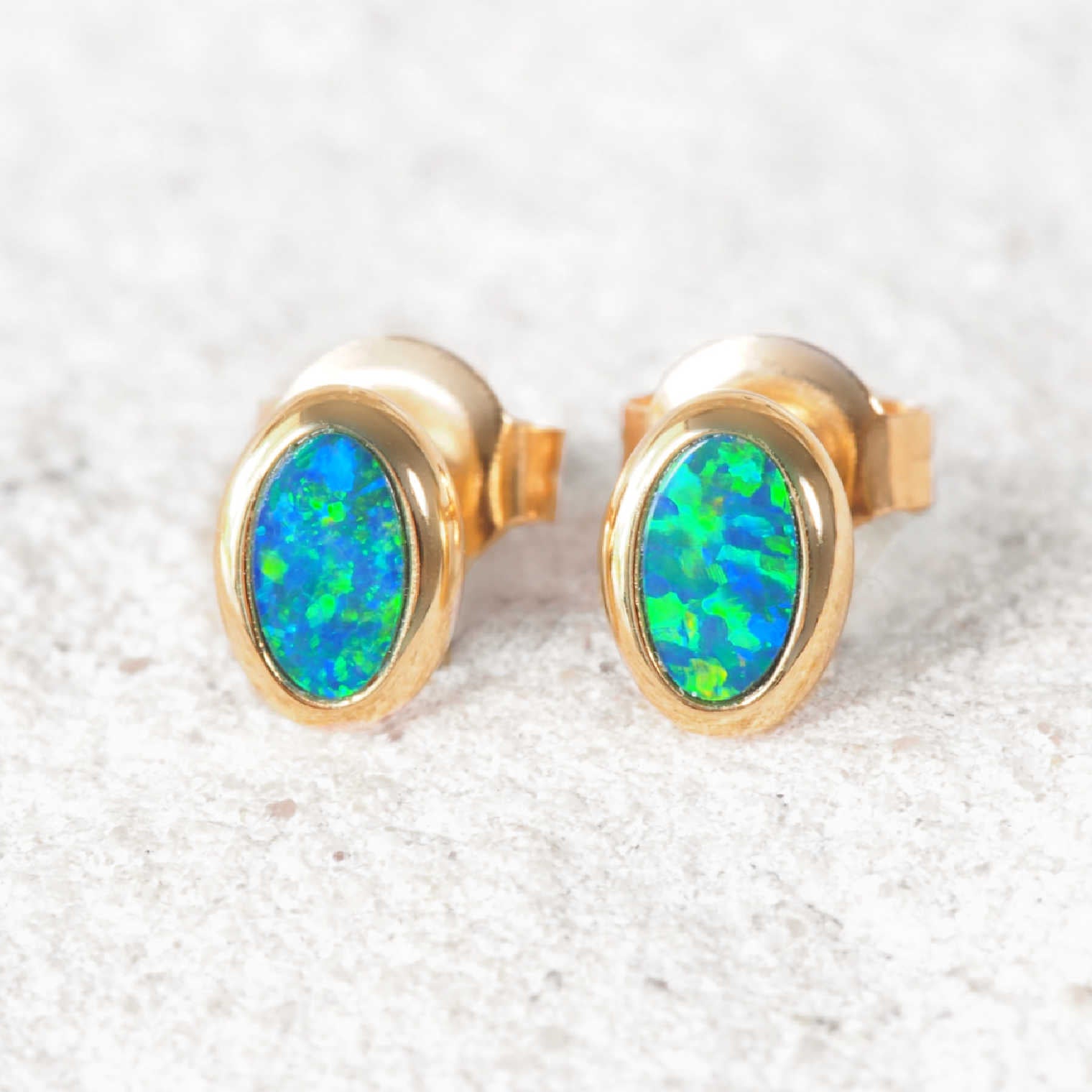 A pair of 14ct gold Australian oval stud earrings with vibrant blue and green opal inlays, displayed on a light textured stone surface.