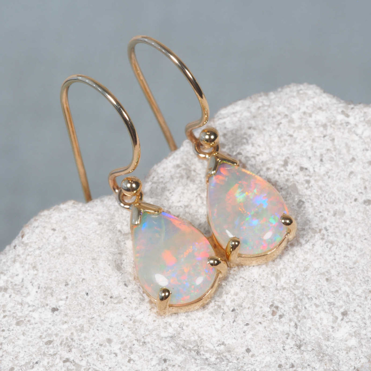 A pair of gold teardrop dangle earrings with iridescent white opal stones, resting on a textured white stone surface against a soft gray background.