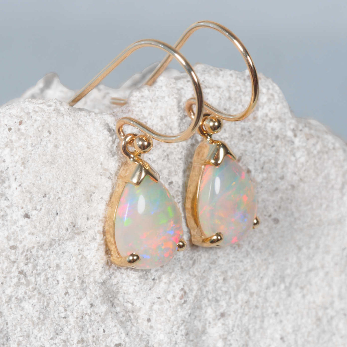 A pair of 14ct yellow gold teardrop dangle earrings with iridescent white opal gemstones, displayed on a textured white stone against a soft gray background.