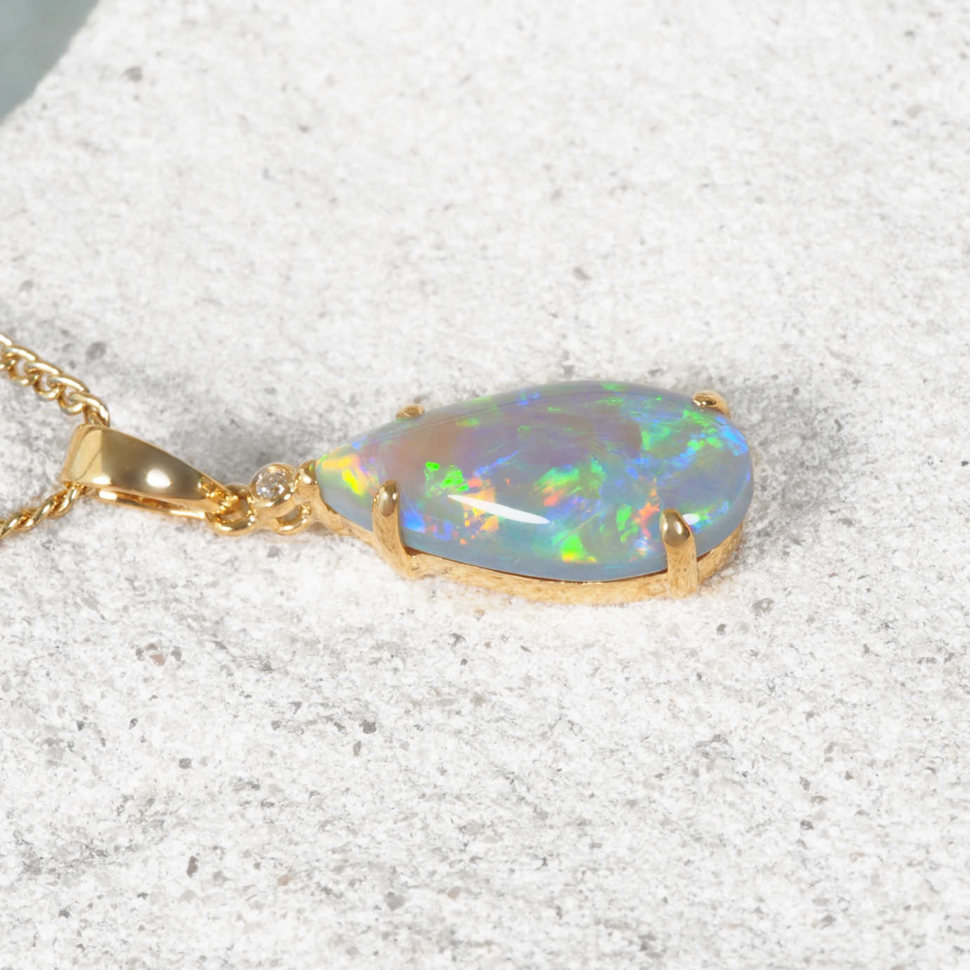 A gold pendant necklace featuring a teardrop-shaped Lightning Ridge opal stone with vibrant iridescent flashes of blue, green, and orange, resting on a textured light stone surface.