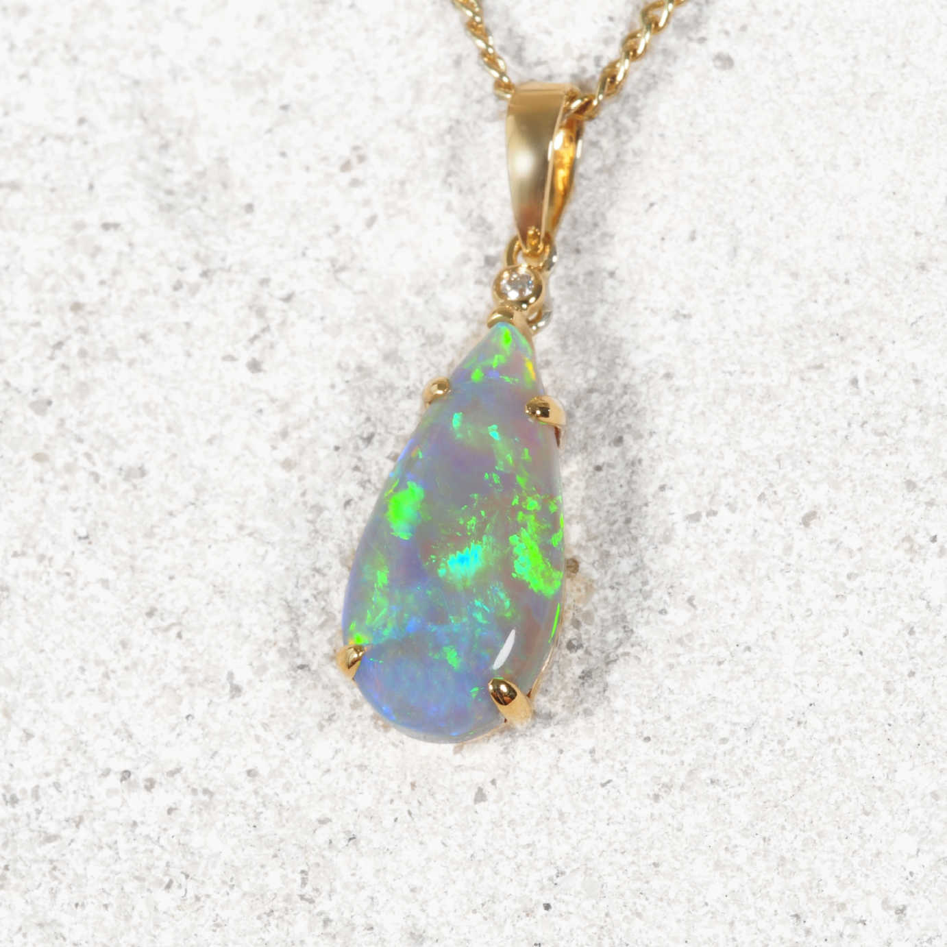 A teardrop-shaped authentic Australian opal pendant necklace in a gold setting on a textured light gray background. The opal displays vibrant green, blue, and white flashes, set in a gold bail with a small clear accent stone.