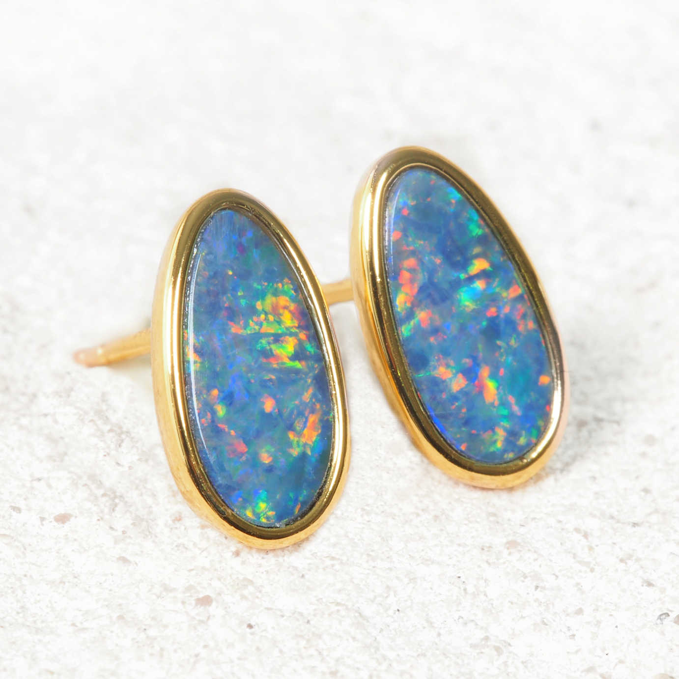 A pair of gold stud earrings with oval-cut blue opal gemstones showing vibrant multicolor flashes, set against a light textured background.
