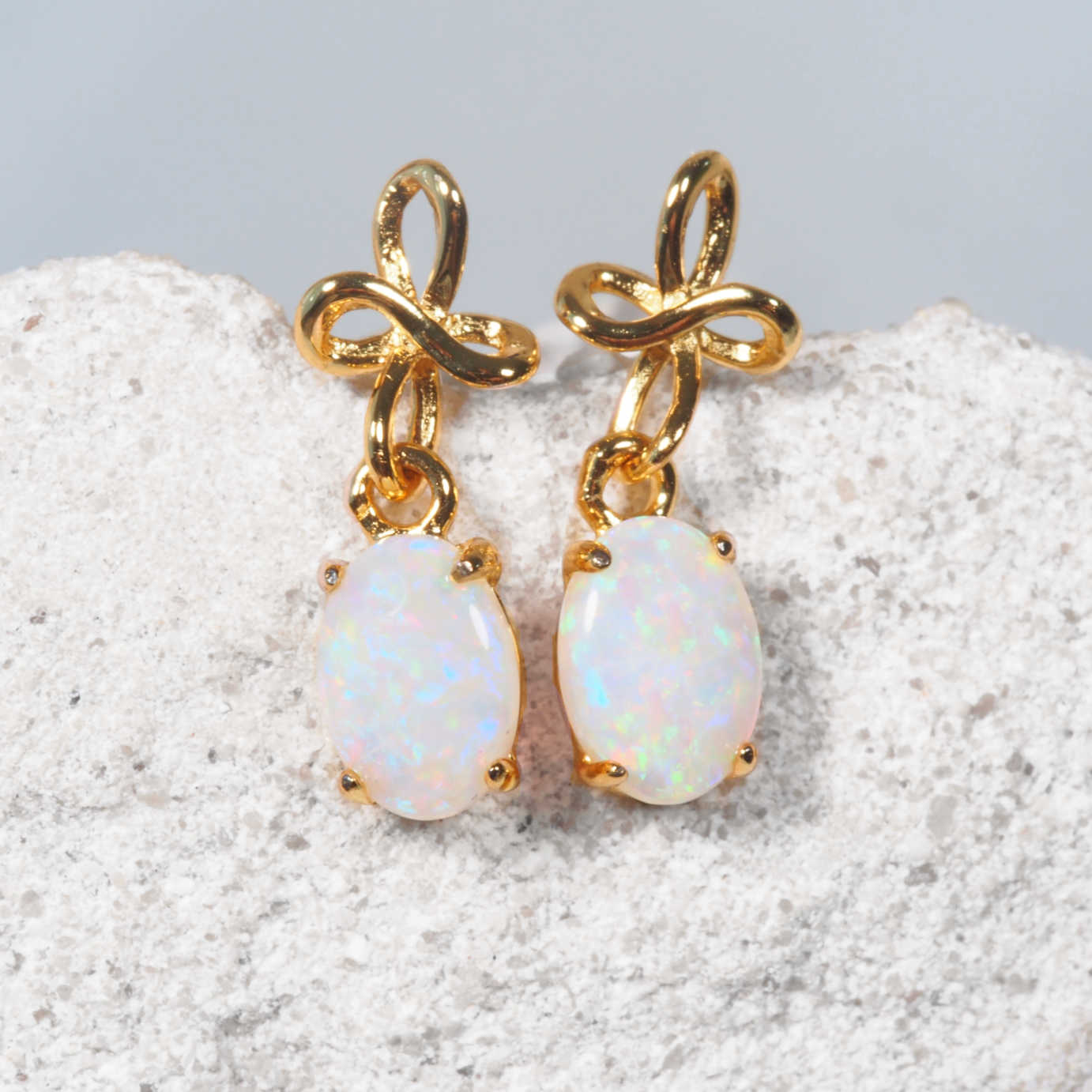 A pair of gold drop Australian opal earrings with oval white opal stones, displayed on a textured light gray stone surface against a light gray background.