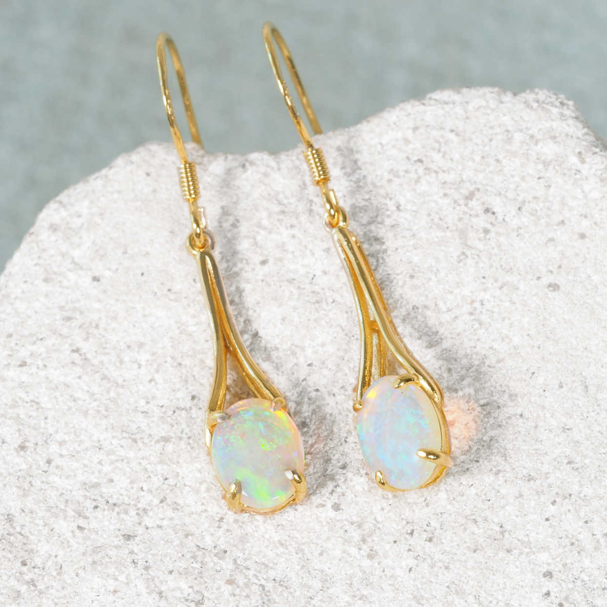 A pair of gold plated silver Australian opal drop earrings with oval iridescent crystal opal stones, displayed on a textured white stone surface against a soft gray background.