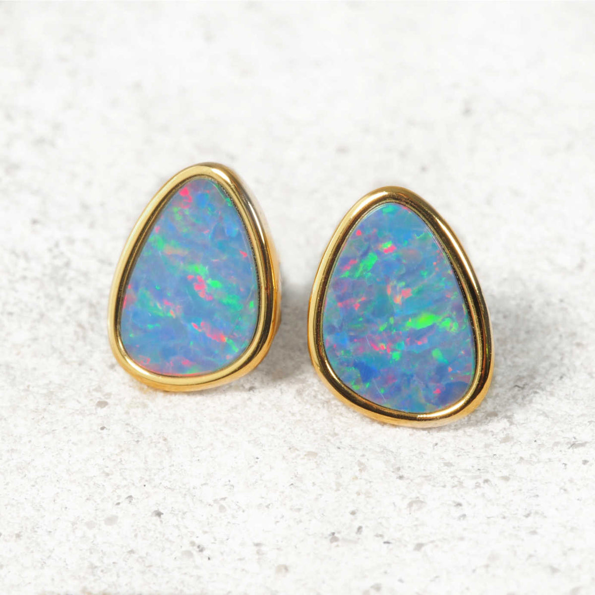 A pair of Australian gold teardrop stud opals earrings with vibrant, iridescent blue, green, and pink opal inlays, set against a light textured stone background.