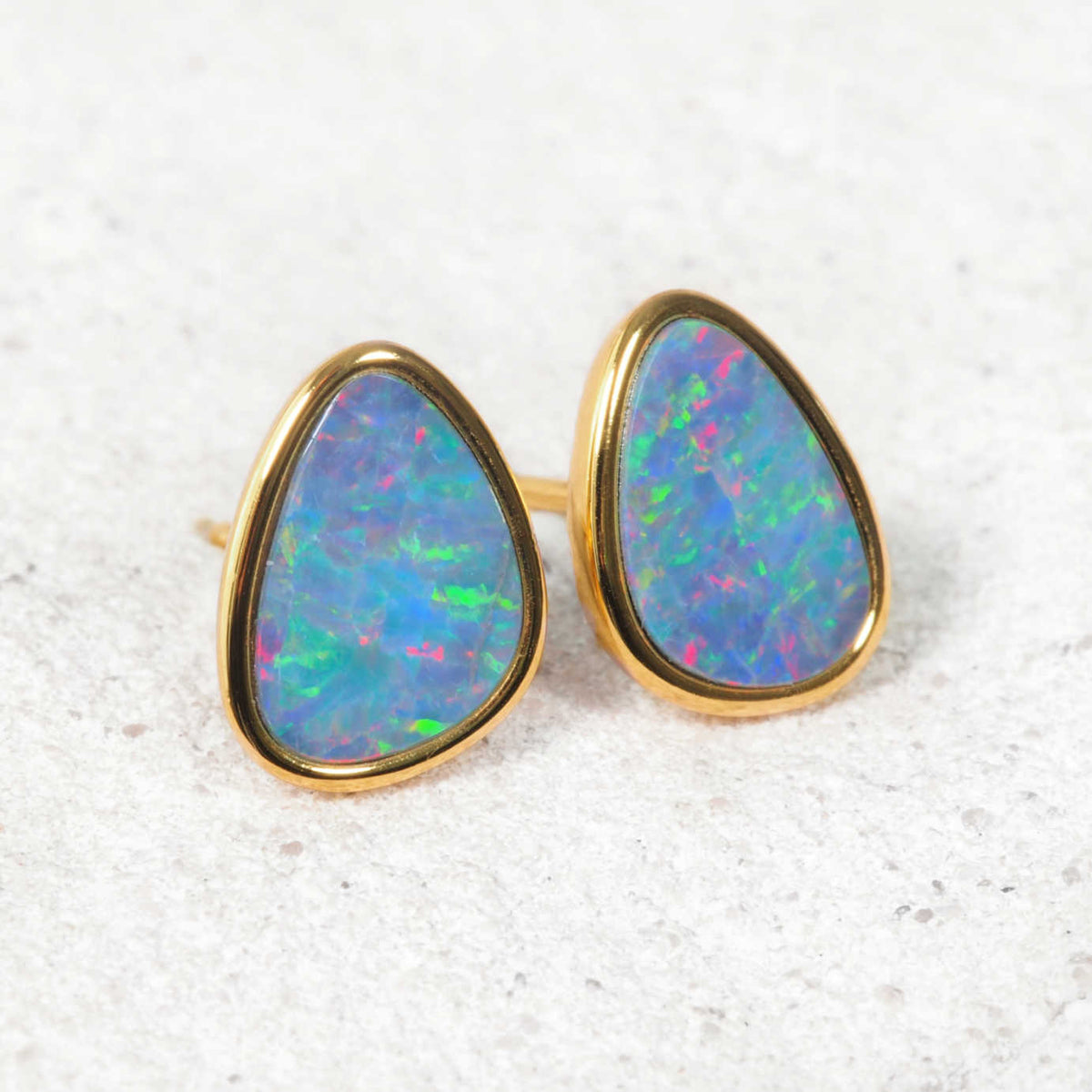 A pair of gold teardrop stud earrings with vibrant, iridescent blue, green, and pink opal inlays, set against a light textured gray background.