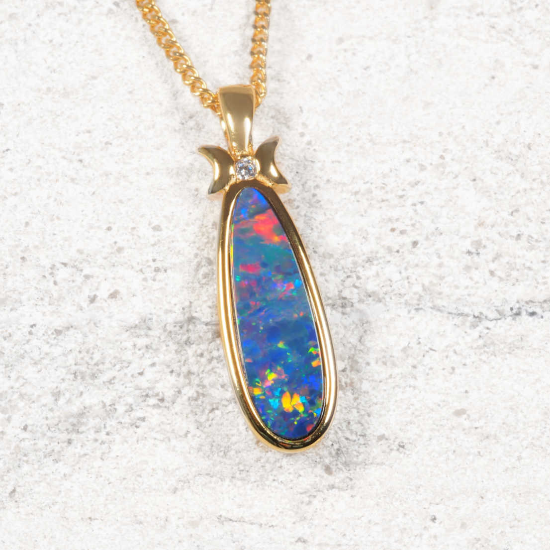 A gold plated silver opal pendant necklace featuring a teardrop-shaped opal stone with vibrant blue, red, and orange play-of-color, set on a textured light gray background.