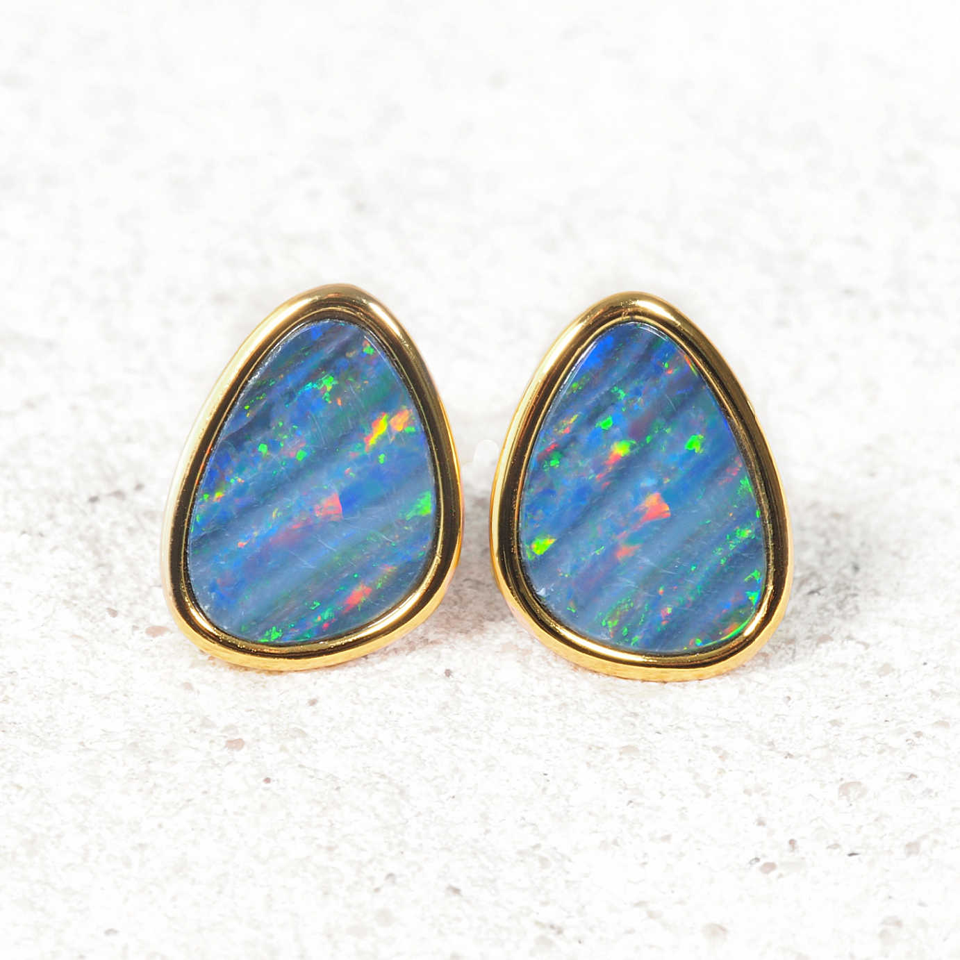 A pair of gold pear-shaped opal stud earrings with vibrant blue opal stones showing flashes of green, orange, and pink, set against a light textured background.