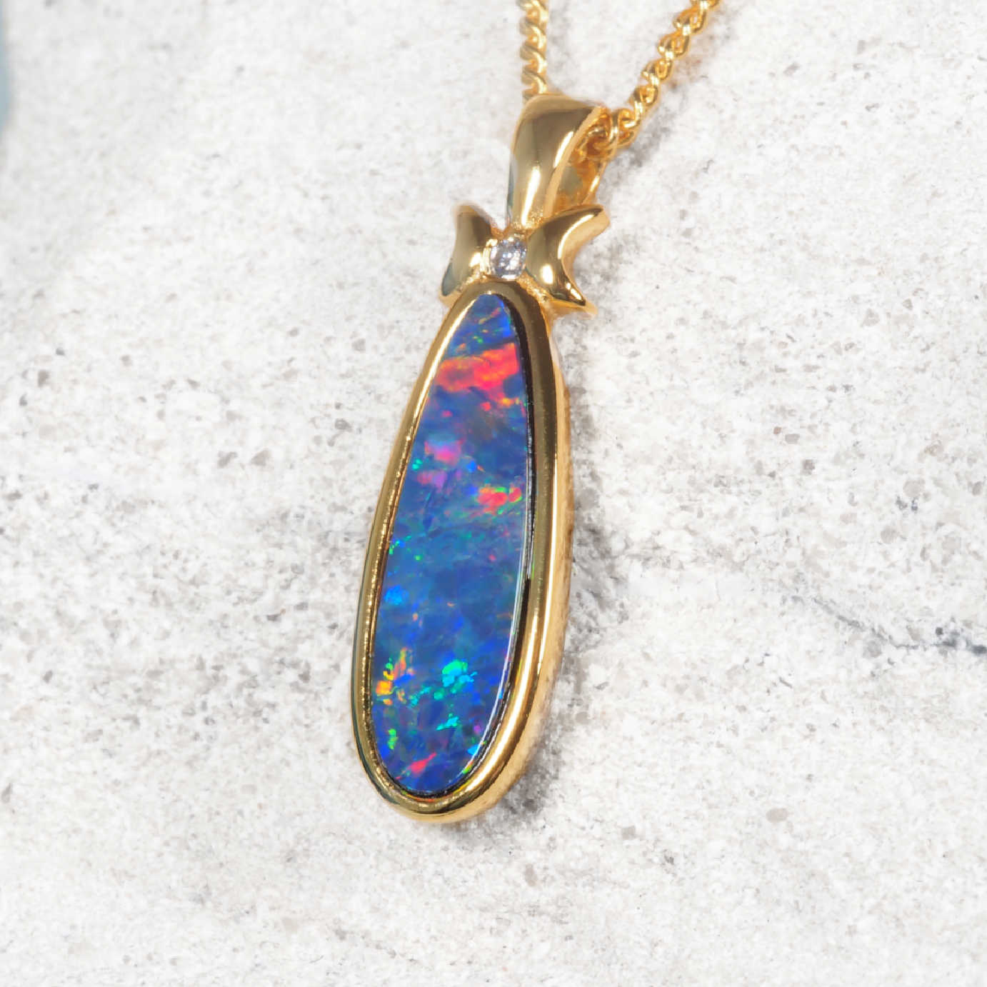 A gold plated silver Australian opal pendant necklace featuring a teardrop-shaped opal stone with vibrant blue, red, and green flashes, set on a textured light gray stone background.