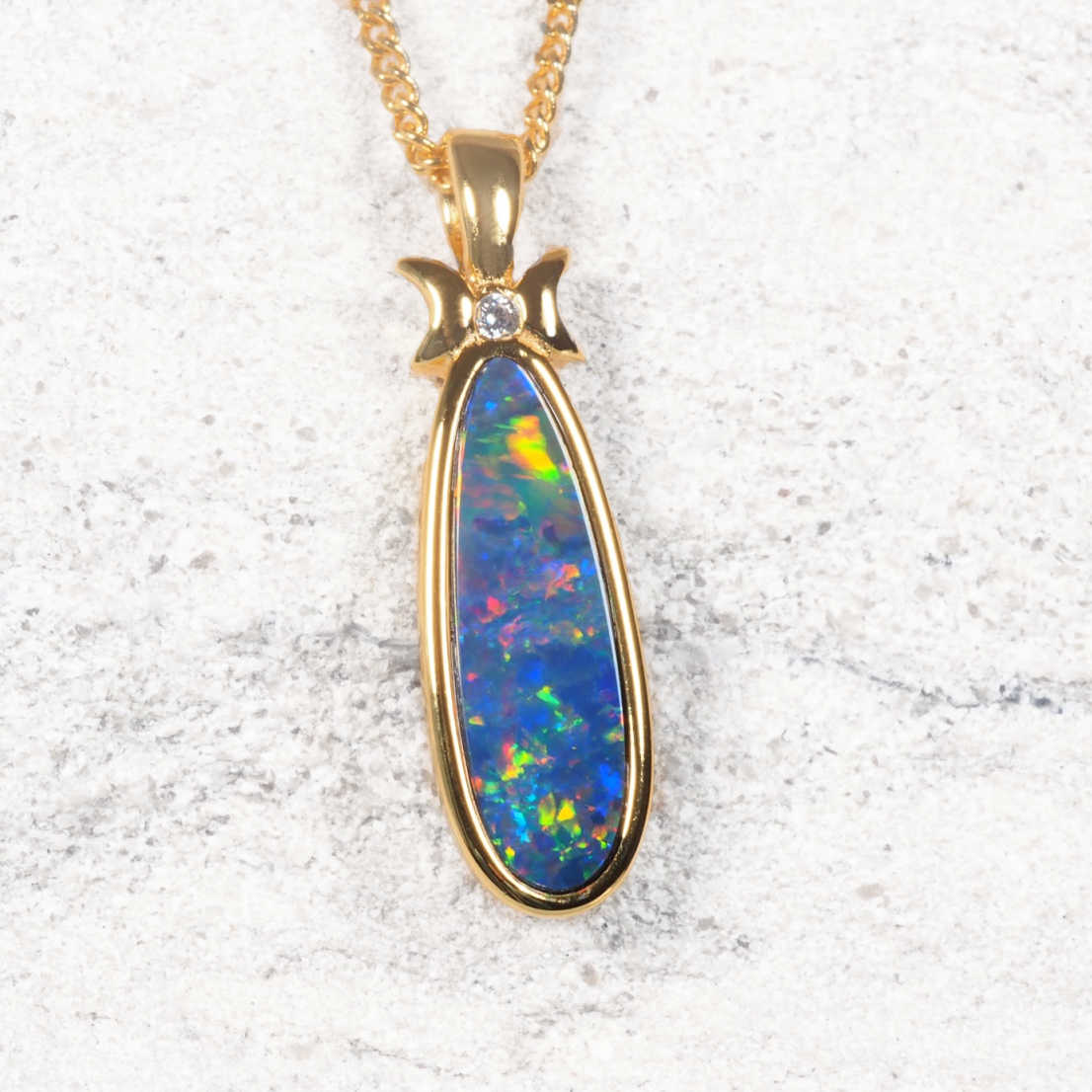 A gold plated silver natural doublet opal pendant necklace featuring a teardrop-shaped opal stone with vibrant blue, green, orange, and red flashes, set in a polished gold bezel with a small clear accent stone at the top, resting on a textured light gray stone surface.