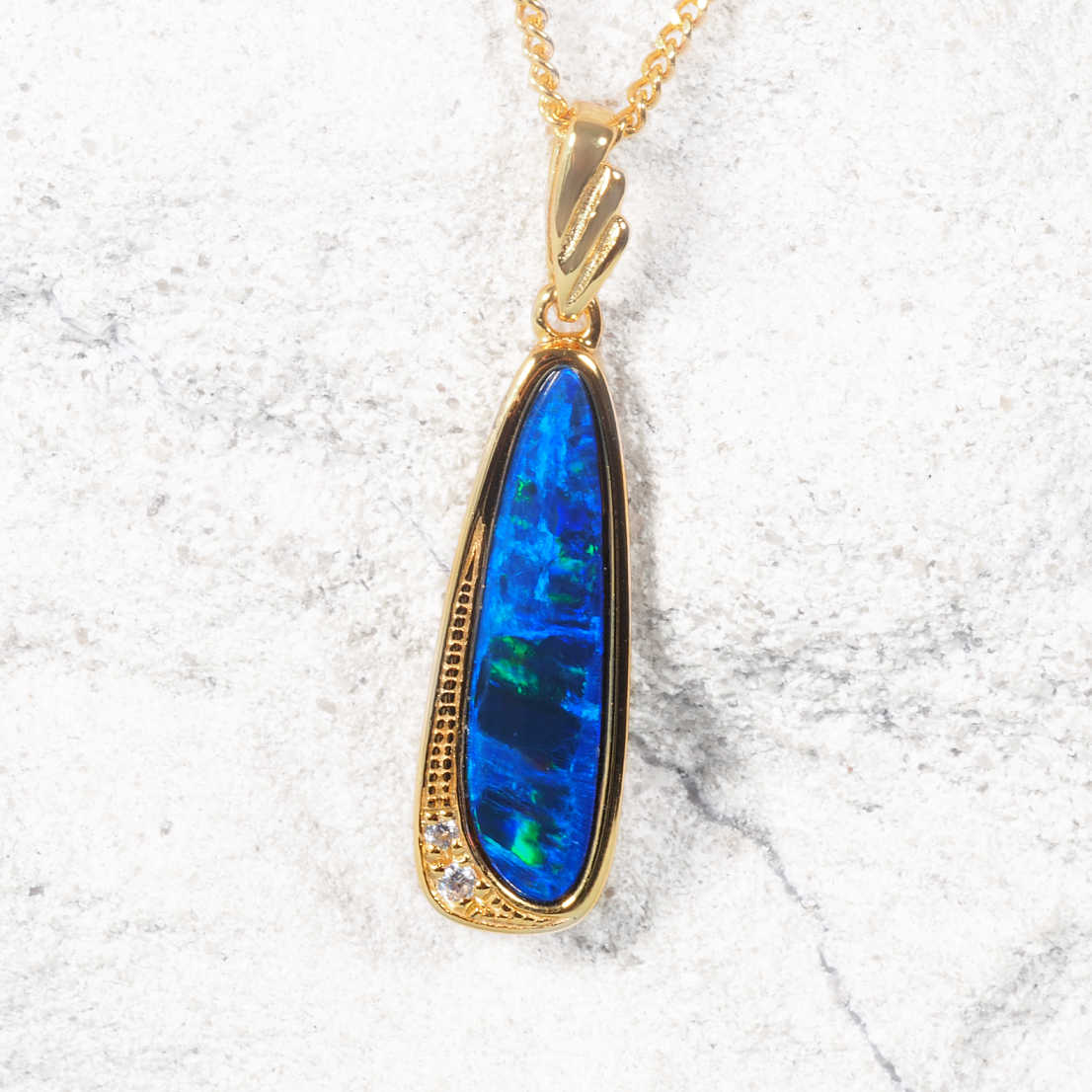 A gold plated silver opal teardrop pendant necklace featuring a vibrant blue opal stone with green flashes, set against a light gray textured stone background.