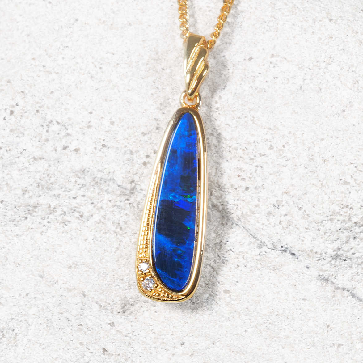 A genuine opal pendant necklace featuring a vibrant blue teardrop-shaped opal stone, set in a gold plated silver bezel with a small clear accent stone on the side, resting on a light gray textured surface.