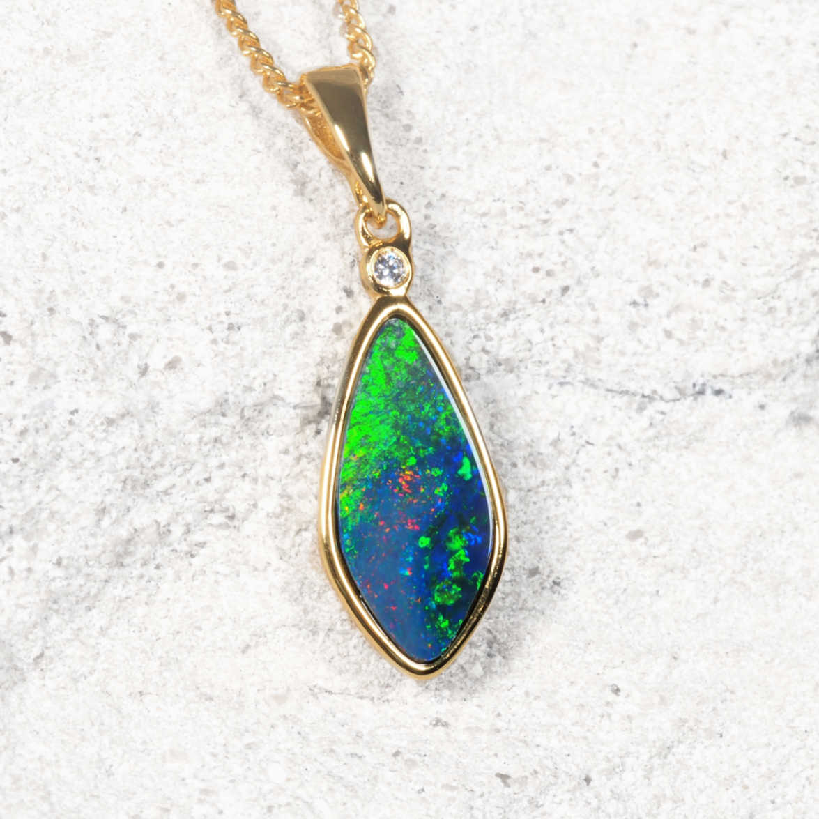 A gold plated silver opal teardrop pendant necklace featuring a vibrant green and blue opal stone with a small clear accent gemstone, displayed on a textured light gray surface.