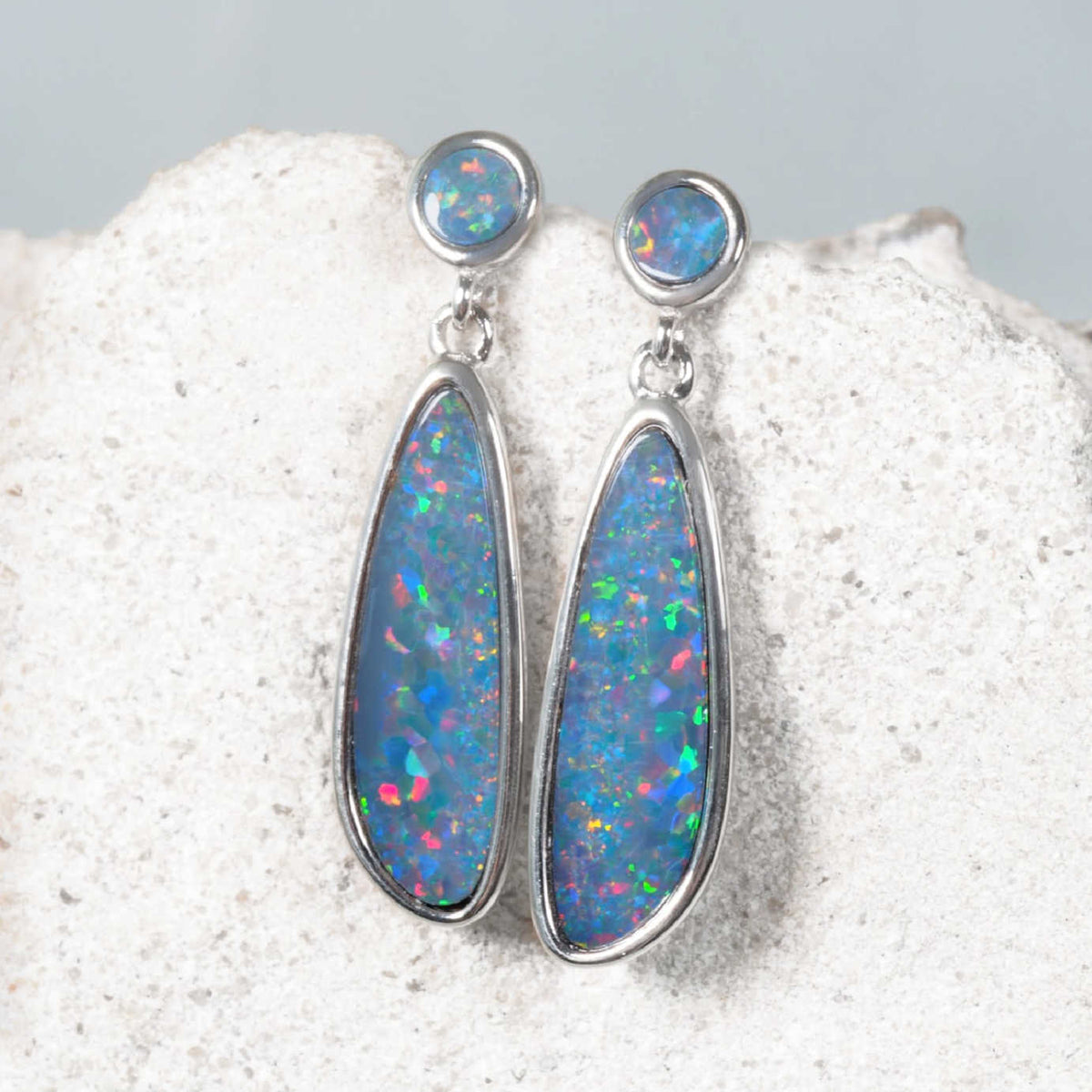 A pair of silver Australian opal dangle earrings with teardrop-shaped blue opal stones and round blue opal studs, displayed on a textured white stone surface.