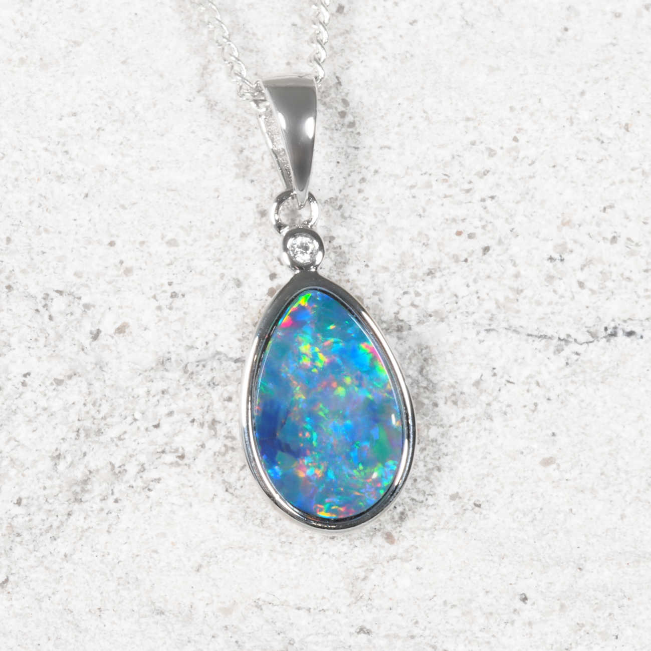A teardrop-shaped doublet opal pendant necklace featuring a vibrant blue and green iridescent opal gemstone in a polished silver bezel setting, suspended from a matching silver chain against a light textured stone background.