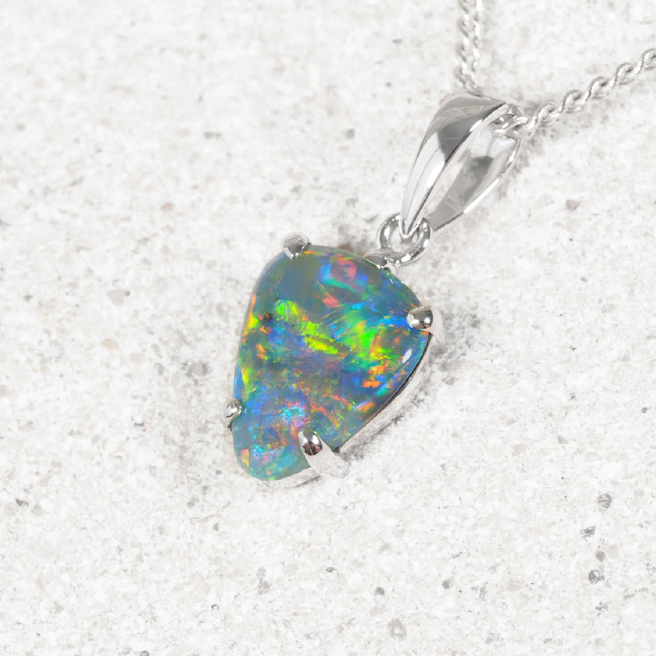 A white gold black opal pendant necklace featuring a teardrop-shaped opal gemstone with vibrant iridescent colors, resting on a light textured surface.