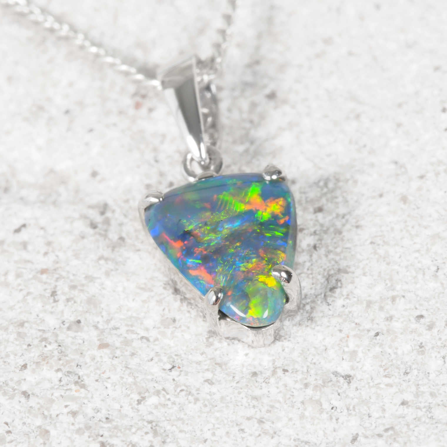 A white gold authentic Australian opal pendant necklace featuring a triangular opal gemstone with vibrant iridescent colors, resting on a textured light gray stone surface.