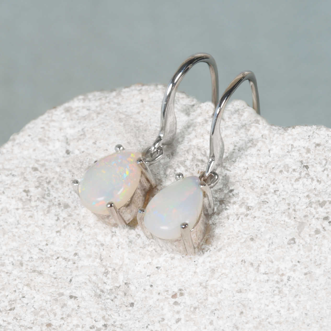 A pair of white gold teardrop-shape authentic Australian opal dangle earrings resting on a textured white stone surface against a soft gray background.