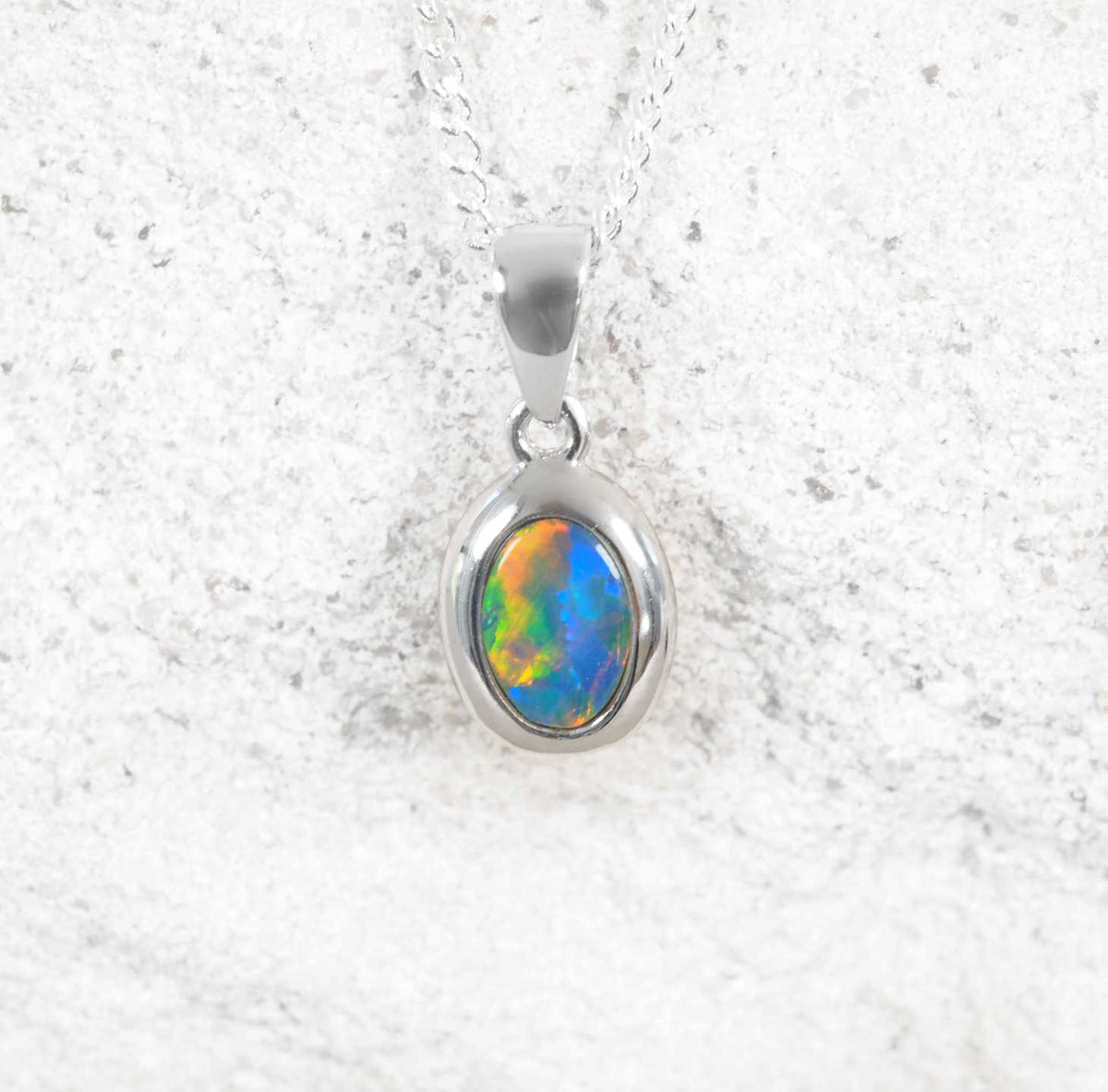 A white gold oval pendant necklace featuring a vibrant, multicolored opal gemstone, displayed on a textured light gray stone surface.