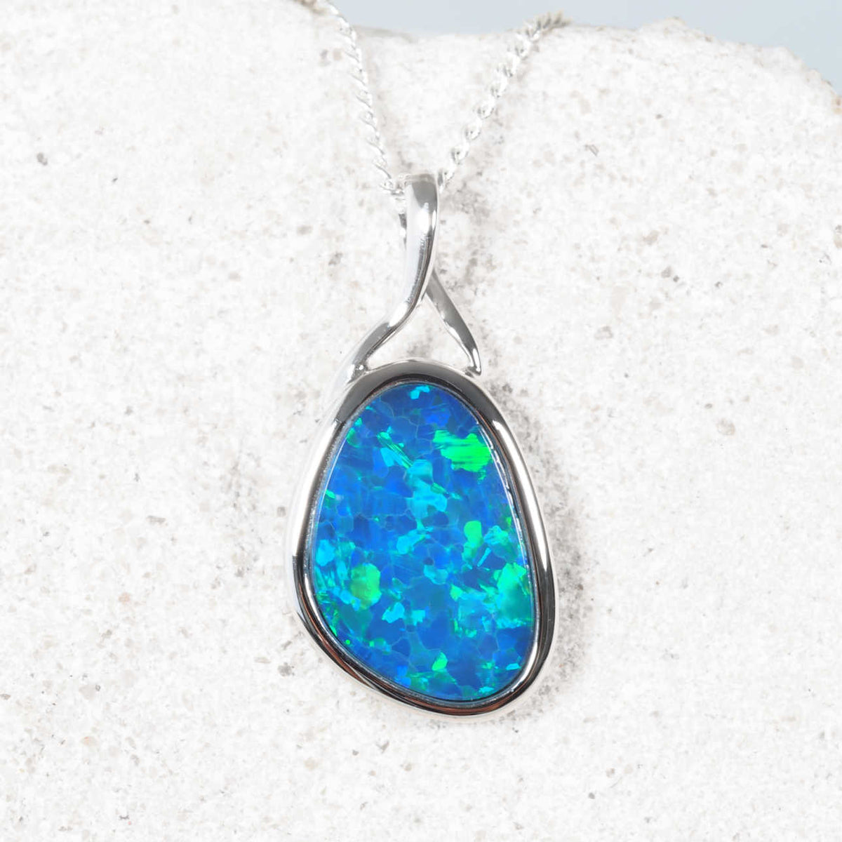 A teardrop-shaped opal pendant necklace set in 14ct white gold, displayed on a textured white stone surface. The opal features vibrant blue and green flashes.