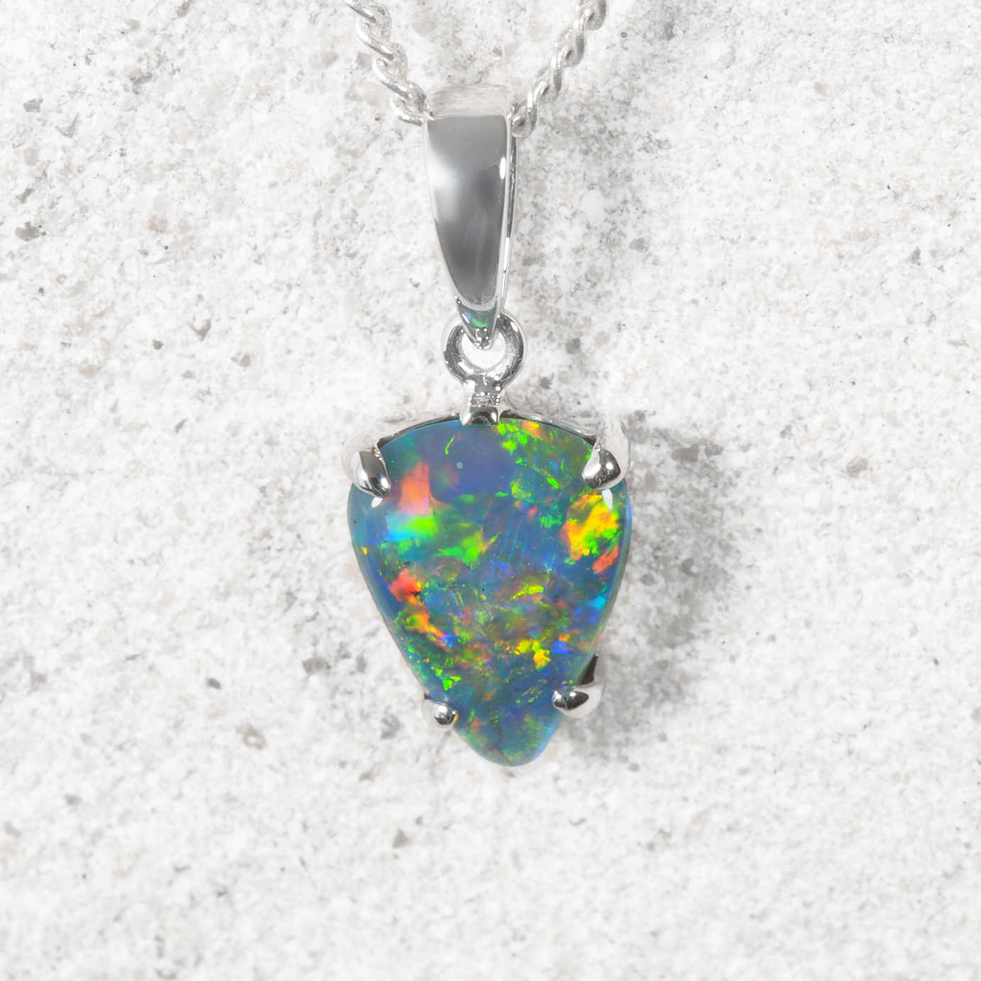 A white gold back opal pendant necklace featuring a teardrop-shaped opal gemstone with vibrant multicolor play-of-color, displayed on a textured light gray stone background.