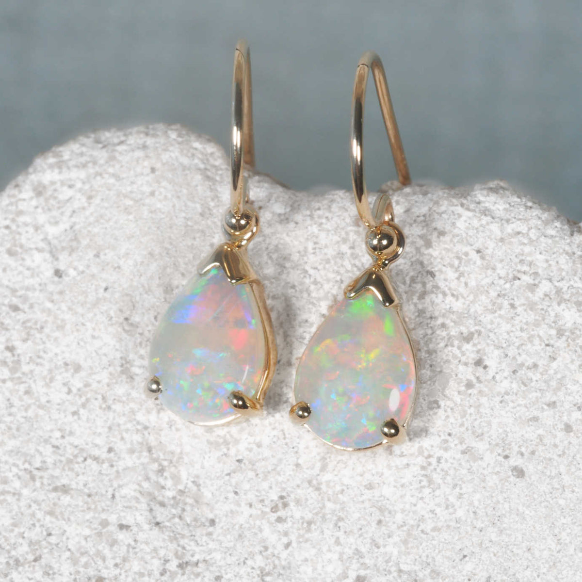 A pair of genuine opal gold teardrop dangle earrings with iridescent white opal gemstones, resting on a textured white stone surface against a soft gray background.