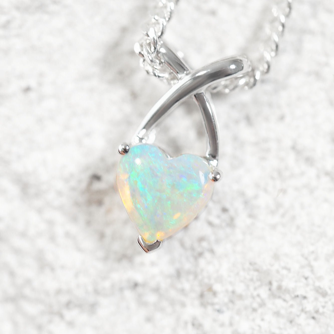 A heart-shaped Australian opal pendant in a polished white gold setting on a silver chain, displayed on a textured light gray surface.
