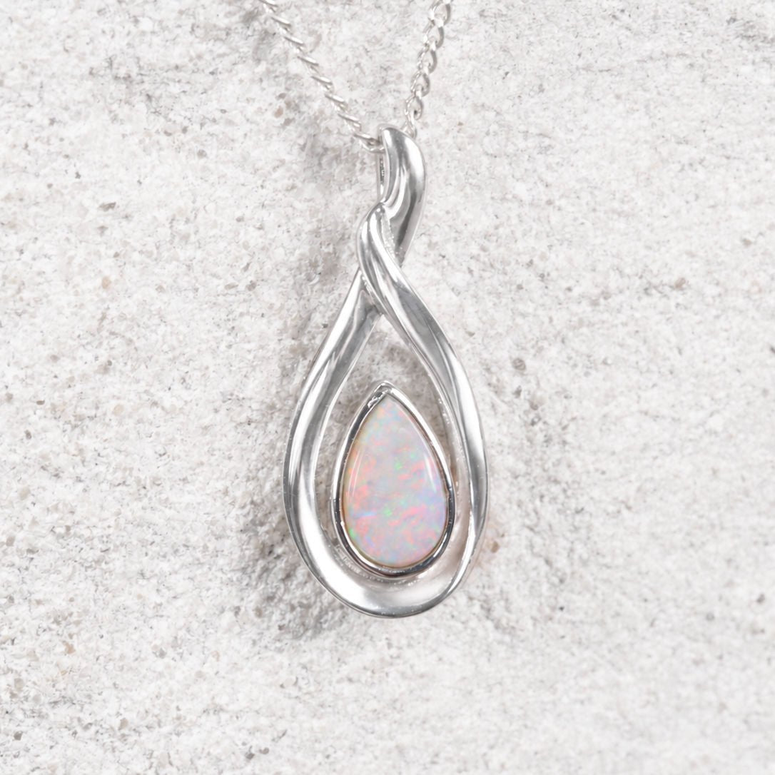 A silver teardrop pendant necklace featuring a central iridescent opal gemstone, displayed on a textured light gray surface.