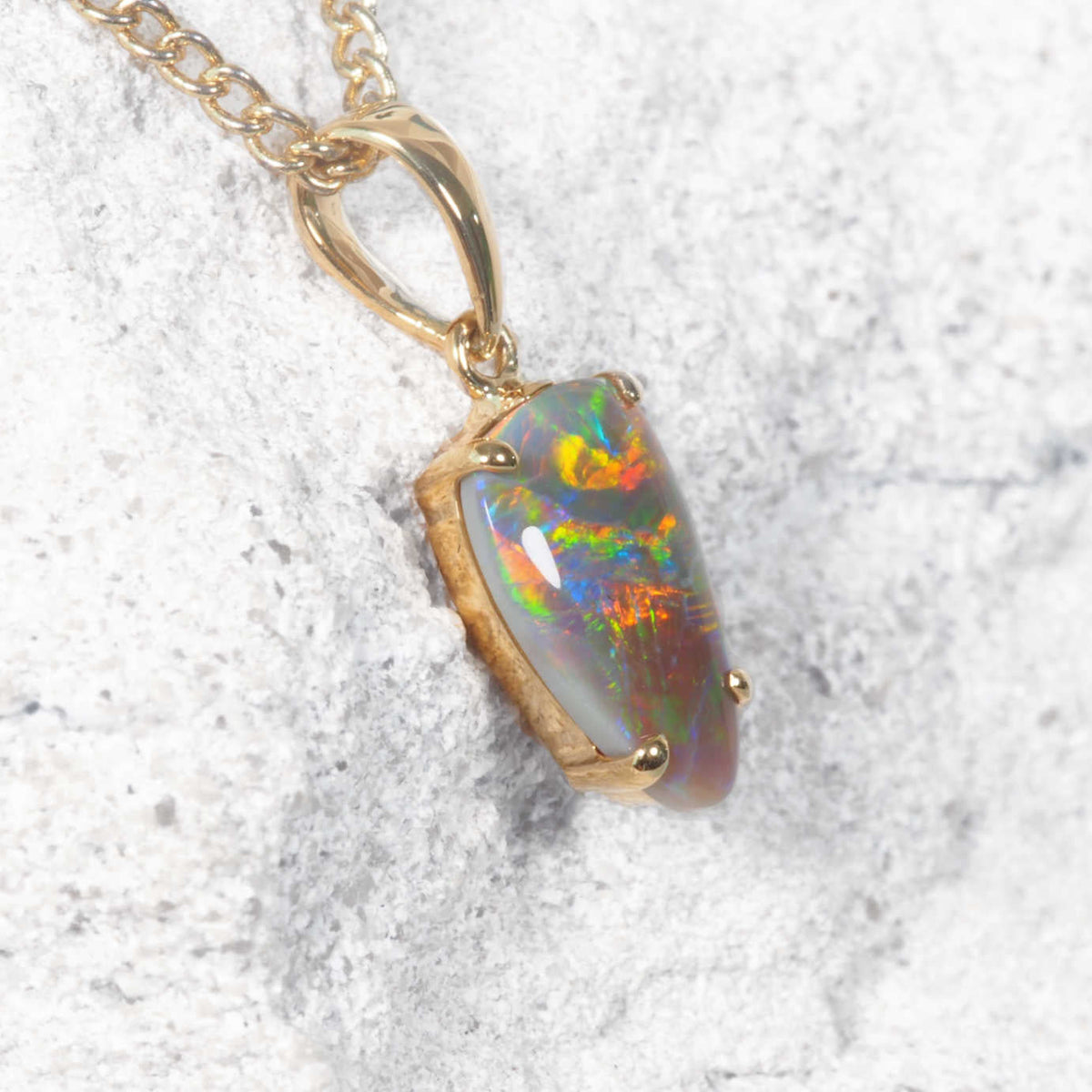 A yellow gold black opal pendant featuring a polished iridescent Australian opal stone with vibrant rainbow play-of-color, set in a gold bezel and hanging from a gold chain against a textured light gray background.
