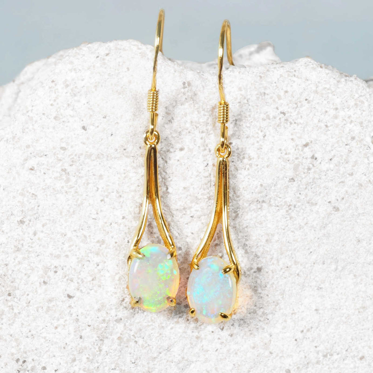 A pair of gold plated silver Australian drop opal earrings with oval green crystal opal stones, displayed on a textured white stone surface.