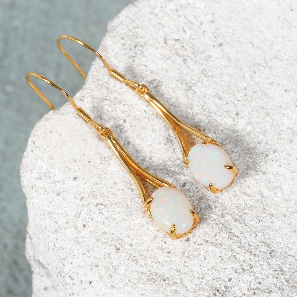 A pair of gold plated silver drop earrings with oval natural Australian white opal stones, displayed on a textured light gray stone surface.