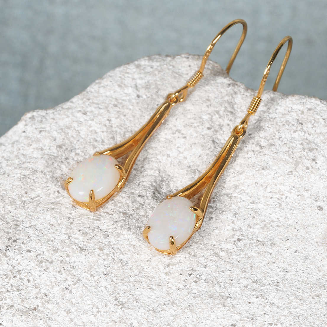 A pair of gold plated silver opal drop earrings with oval white Australian opal stones, displayed on a textured light gray stone surface against a soft gray background.
