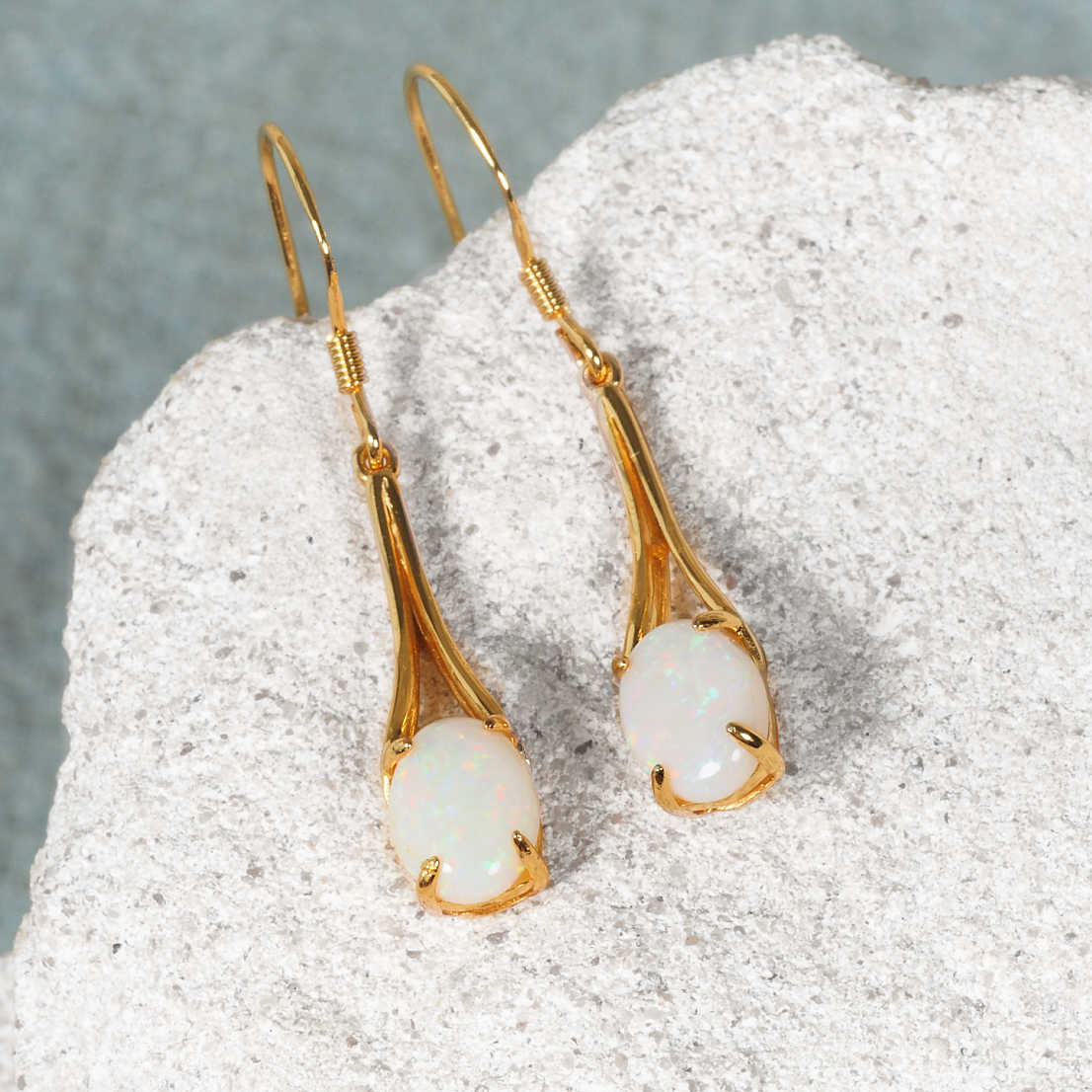 A pair of Australian gold plated silver opal drop earrings with oval Coober Pedy white opal stones, displayed on a textured light gray stone surface.