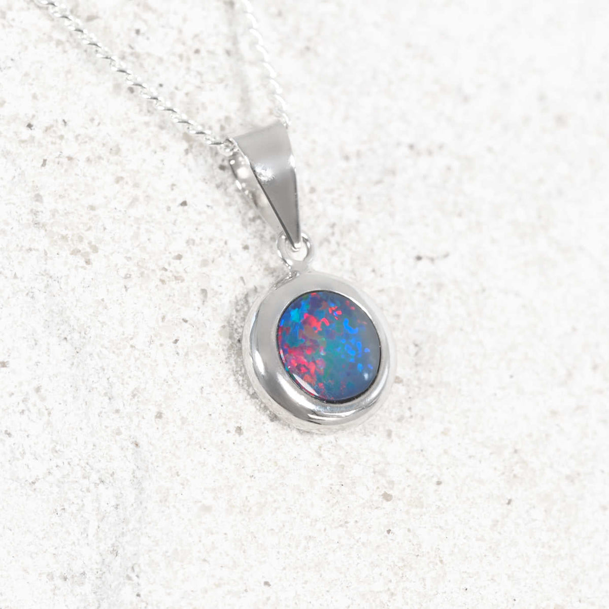 A 14ct white gold doublet opal pendant necklace featuring an oval-cut opal gemstone with vibrant blue, pink, and green flashes, set in a polished silver bezel on a matching silver chain, displayed on a light textured stone surface.