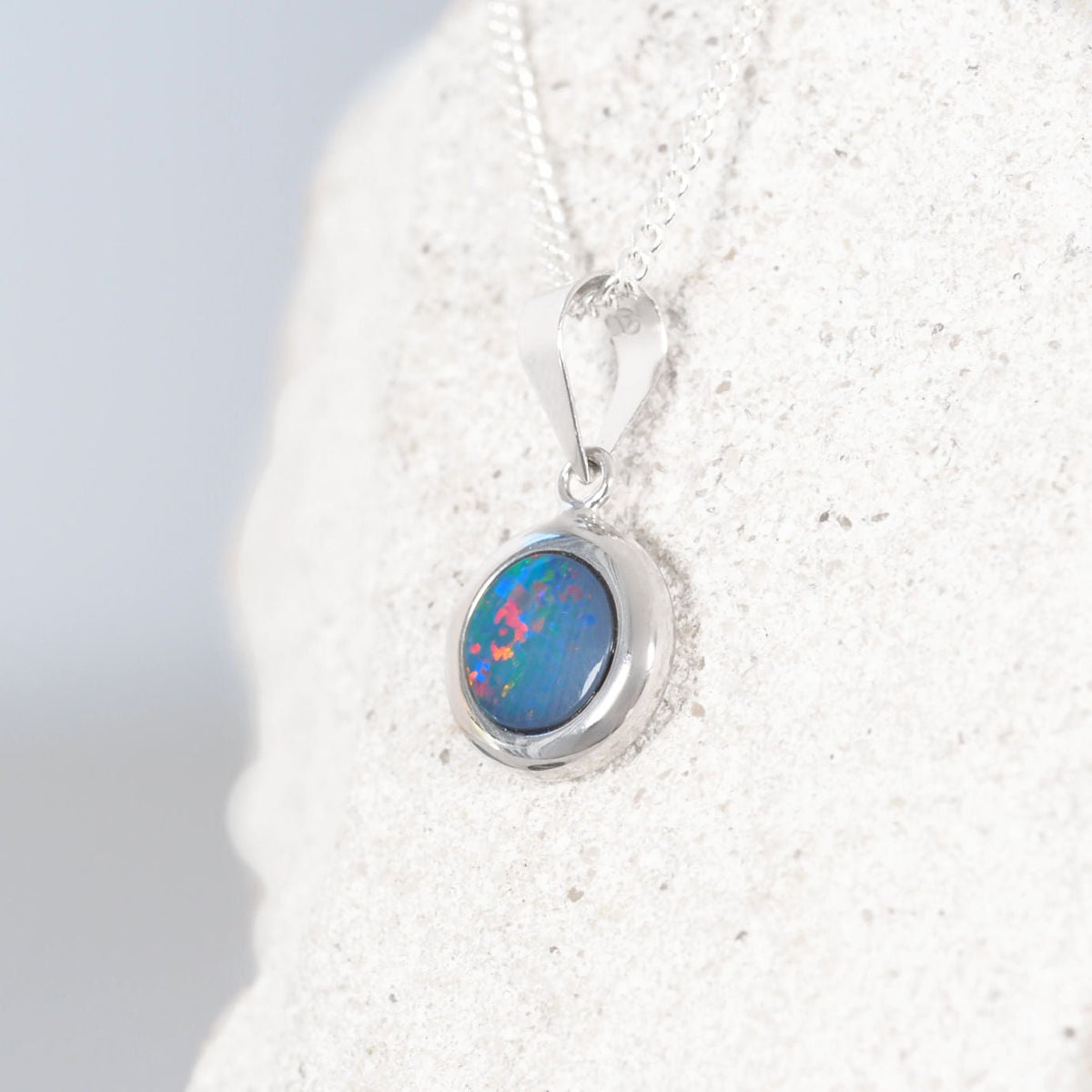 A 14ct white gold opal pendant featuring an oval blue opal with vibrant pink and green flashes, displayed on a textured white stone stand.