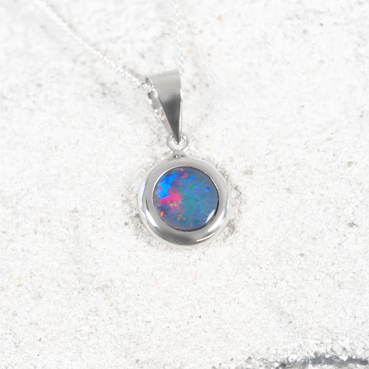 A white gold opal pendant necklace featuring a round bezel-set opal gemstone with vibrant blue, pink, and green flashes, resting on a textured white sand background.