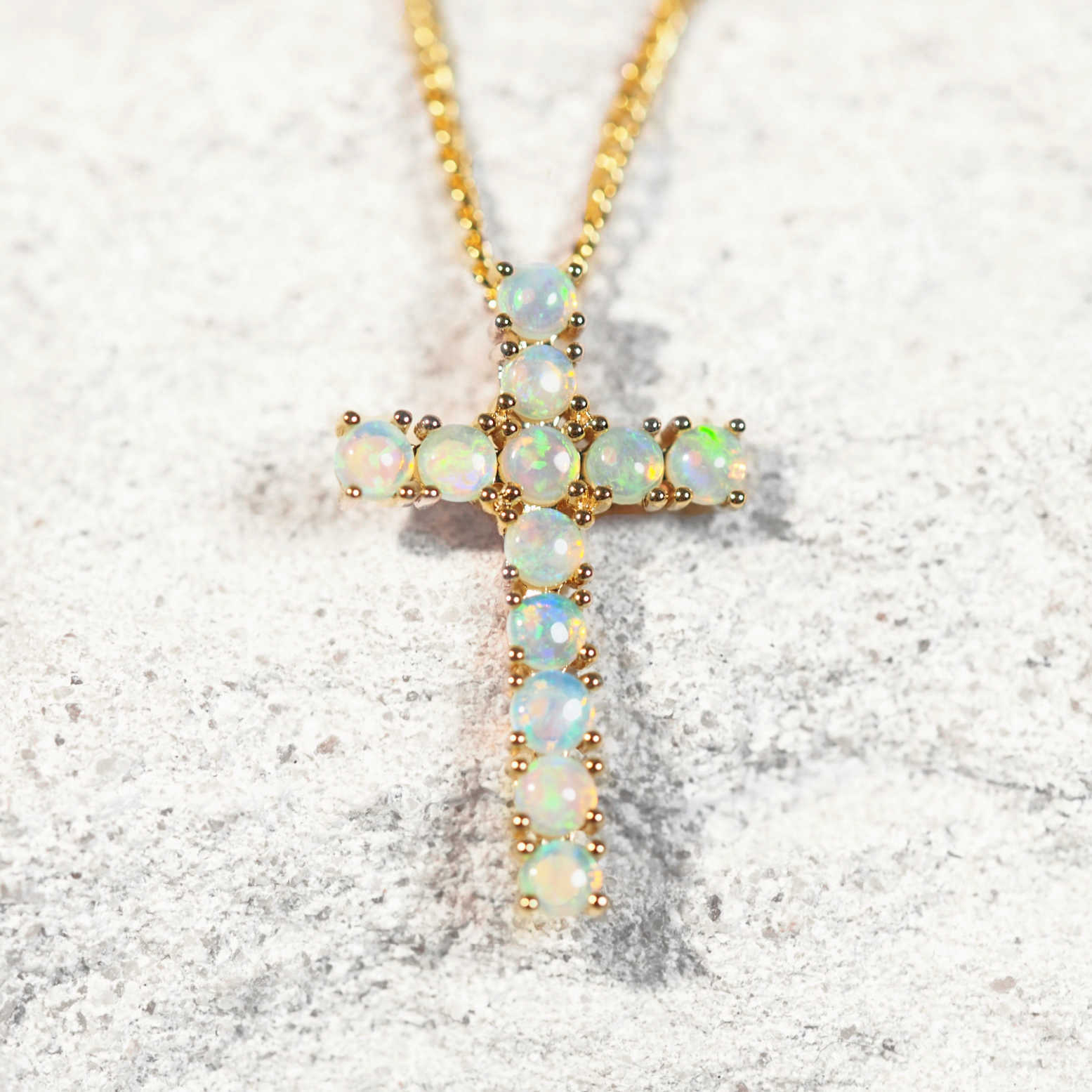 A gold cross opal pendant necklace featuring round iridescent opal stones, displayed on a textured light gray surface.