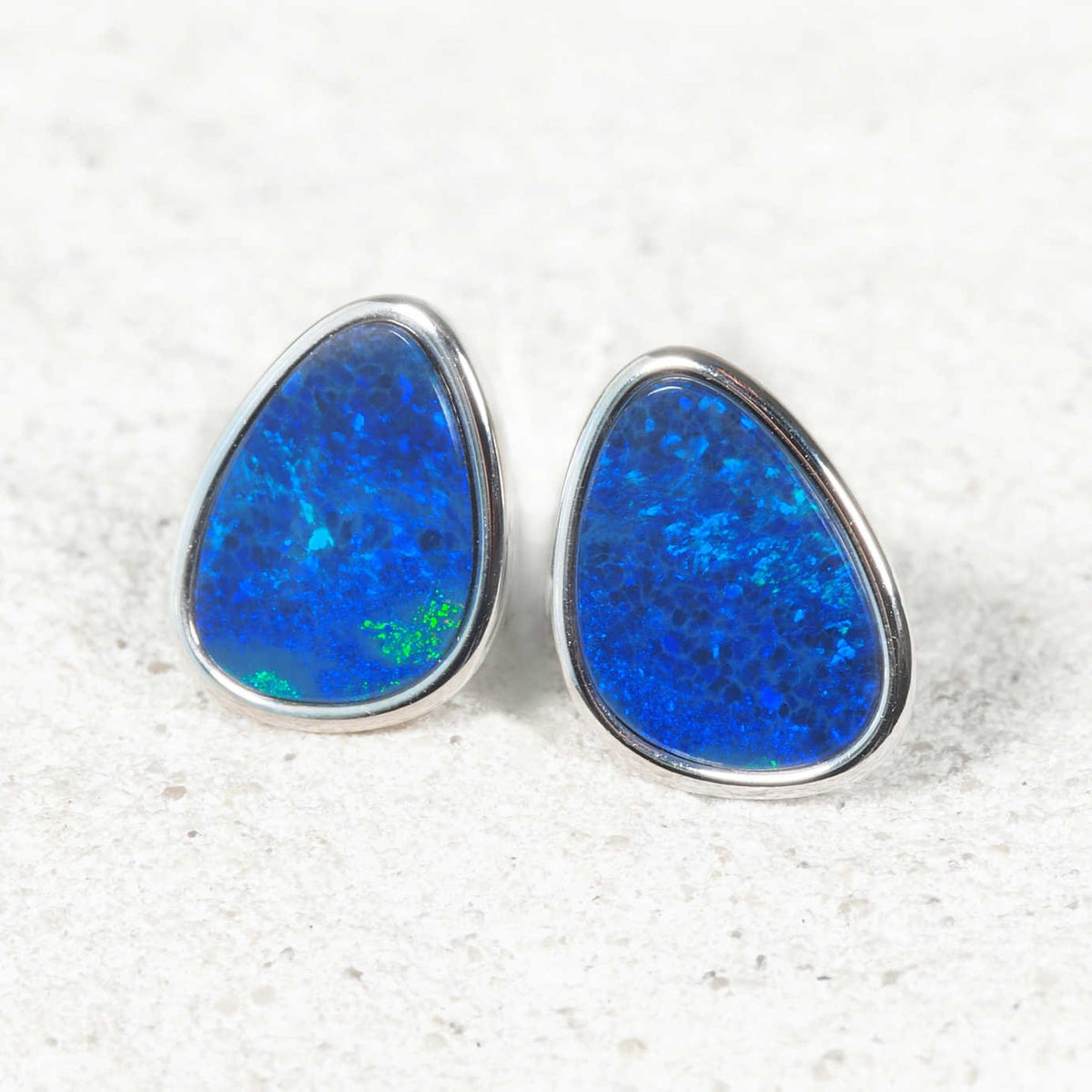 A pair of teardrop-shaped blue opal stud earrings with silver bezel settings, resting on a light textured surface.