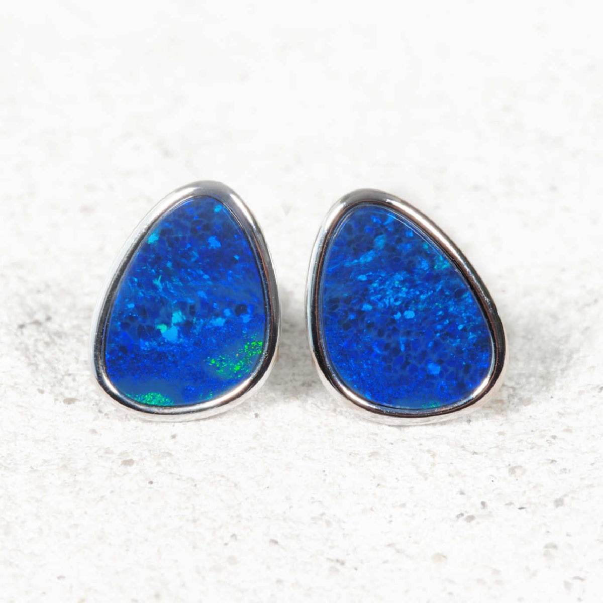 A pair of teardrop-shaped opal stud earrings featuring vibrant blue opal inlays set in a polished silver-tone metal, displayed on a light textured surface.