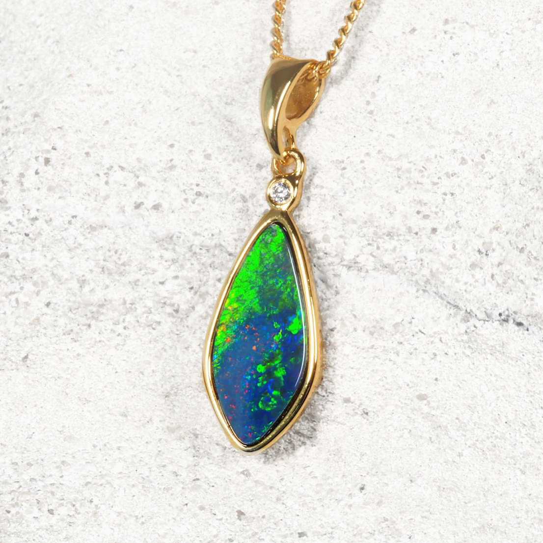 A genuine Australian opal gold plated silver pendant necklace featuring a teardrop-shaped opal stone with vibrant green, blue, and red play-of-color, accented by a small clear stone and a gold chain, set against a light textured background.