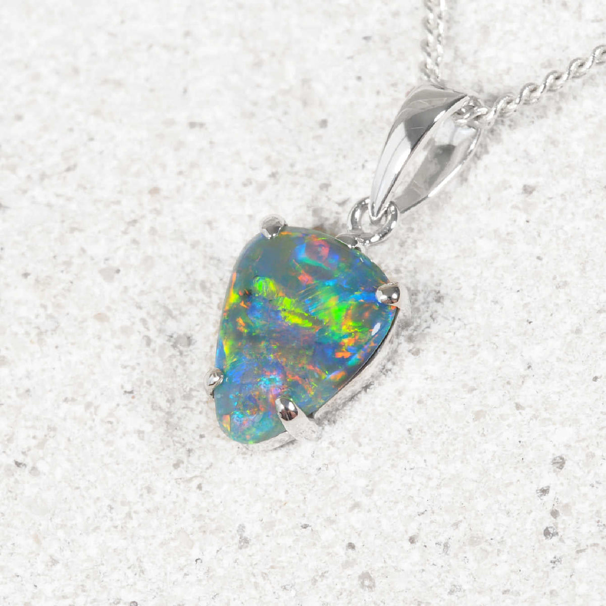 A white gold black opal pendant necklace featuring a teardrop-shaped opal gemstone with vibrant iridescent colors, resting on a light textured surface.