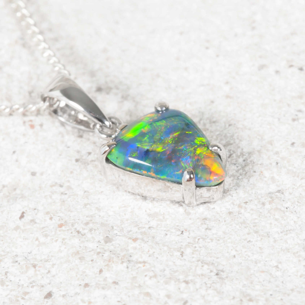 A white gold black opal pendant necklace featuring a triangular opal gemstone with vibrant green, blue, and orange play-of-color, resting on a textured light gray surface.