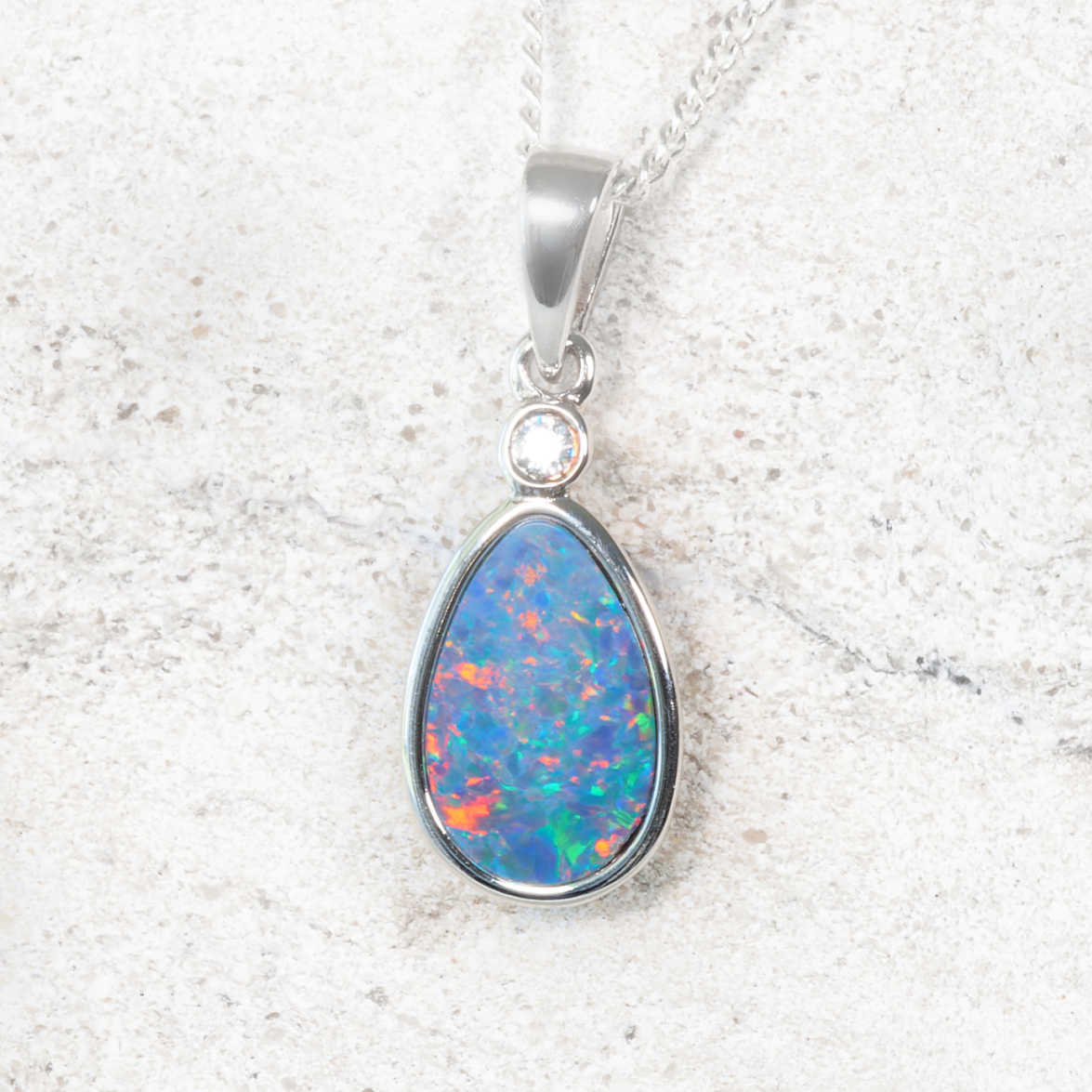 A sterling silver opal teardrop pendant necklace featuring a vibrant blue, green, and orange Australian opal gemstone, set in a polished silver bezel with a round crystal stone above it, resting on a light textured stone surface.