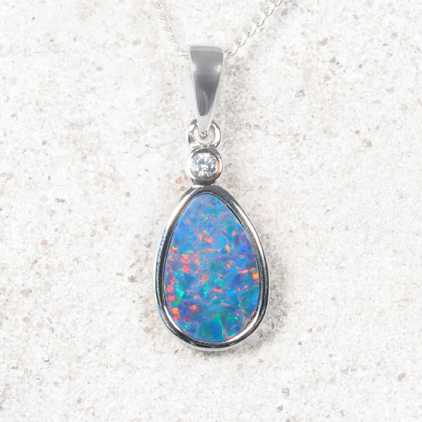 A silver teardrop pendant necklace featuring a vibrant blue, green, and orange opal stone, set in a polished silver bezel with a small clear accent stone, resting on a light textured stone surface.