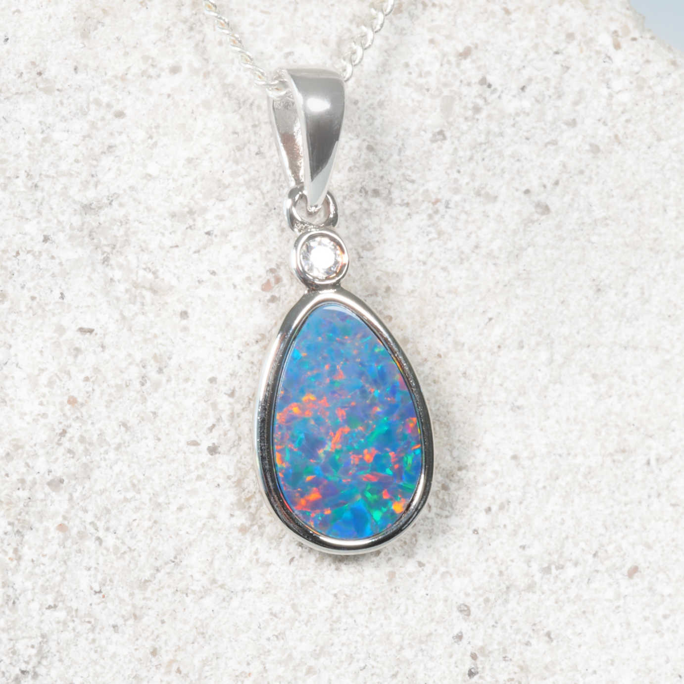 A silver teardrop pendant necklace featuring a vibrant blue opal with flashes of orange and green, set in a polished silver bezel with a small clear accent stone, resting on a textured light gray stone surface.
